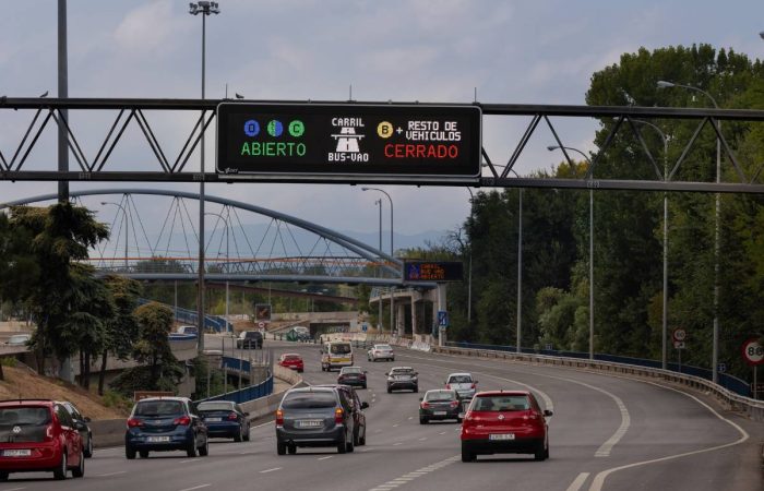 Señalización vial inteligente en autopista urbana que indica carril BUS-VAO abierto para vehículos autorizados y cerrado para el resto, con panel luminoso LED sobre la carretera y tráfico circulando bajo un puente peatonal.