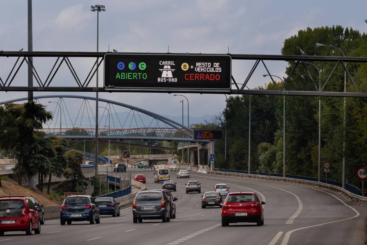 Señalización vial inteligente en autopista urbana que indica carril BUS-VAO abierto para vehículos autorizados y cerrado para el resto, con panel luminoso LED sobre la carretera y tráfico circulando bajo un puente peatonal.